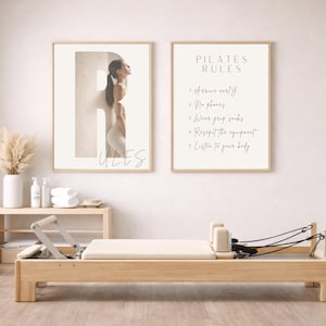 Könnte beinhalten: Zwei gerahmte Drucke und ein Pilates-Reformer. Ein Druck zeigt die Silhouette einer Frau in einem großen Buchstaben, darunter das Wort „RULES“. Der zweite Druck zeigt „PILATES RULES“ und Anweisungen. Der Reformer ist aus Holz.