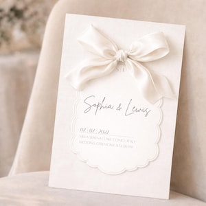 Könnte beinhalten: Elfenbeinfarbene Hochzeitseinladung mit einer Satinschleife. Die Einladung zeigt die Namen "Sophia & Lewis" und das Datum "07.07.2027". Der Text ist in einer zarten, eleganten Schriftart gehalten. Creme Hintergrund.
