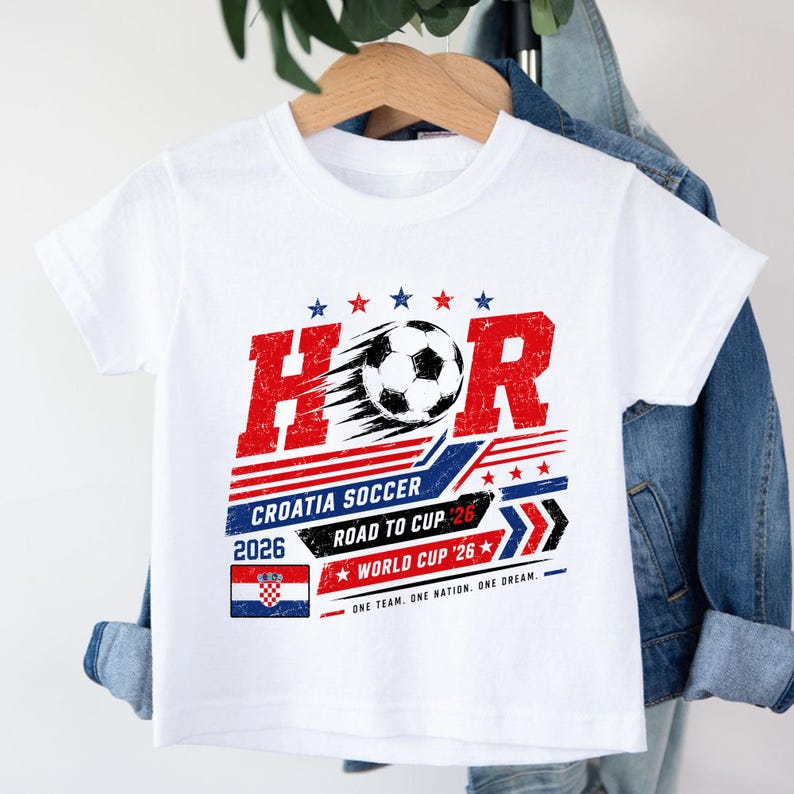 Może przedstawiać: Biały t-shirt z kr&oacute;tkim rękawem z grafiką przedstawiającą piłkę nożną, napis HOR, CROATIA SOCCER, ROAD TO CUP 26, WORLD CUP 26 oraz flagę Chorwacji. Projekt wykorzystuje czerwone, białe, niebieskie i czarne kolory z efektem postarzenia.