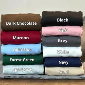 Może przedstawiać: Stos złożonych koszulek w r&oacute;żnych kolorach, w tym Dark Chocolate, Maroon, Light Blue, Forest Green, Sport Grey, Black, Pink, Grey, White, Navy i Sand. Każda koszulka jest oznaczona odpowiednią nazwą koloru w białym tekście.