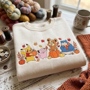 Peut inclure: Un sweat-shirt beige à col rond orné d'une broderie représentant quatre personnages de dessin animé : un Pikachu jaune tenant une citrouille, un Mélofée rose, un Teddiursa marron assis sur une citrouille et un Ronflex bleu portant une écharpe.