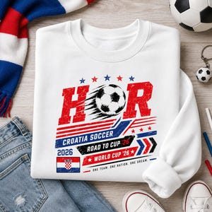 Może przedstawiać: Biała bluza z okrągłym dekoltem z nadrukiem graficznym zawierającym napis HOR, piłkę nożną oraz Croatia Soccer Road to Cup 26 World Cup 26. Wz&oacute;r zawiera czerwone, białe i niebieskie kolory, gwiazdy, paski oraz emblemat flagi chorwackiej.