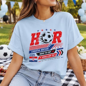 Może przedstawiać: Jasnoniebieski t-shirt z grafiką przedstawiającą napis HOR, piłkę nożną oraz słowa CROATIA SOCCER, ROAD TO CUP 26 i WORLD CUP 26. Wz&oacute;r zawiera małą chorwacką flagę, czerwone i niebieskie paski oraz gwiazdki w stylu postarzanym.
