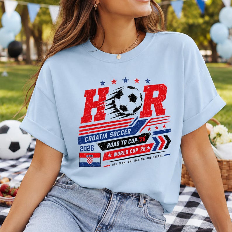 Może przedstawiać: Jasnoniebieski t-shirt z grafiką przedstawiającą napis HOR, piłkę nożną oraz słowa CROATIA SOCCER, ROAD TO CUP 26 i WORLD CUP 26. Wz&oacute;r zawiera małą chorwacką flagę, czerwone i niebieskie paski oraz gwiazdki w stylu postarzanym.