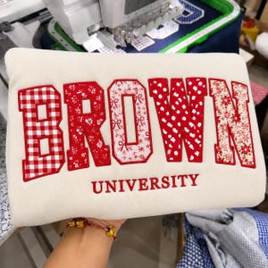 May include: A cream-coloured sweatshirt featuring the word BROWN in large, red-bordered appliqué letters, each with a unique red and white pattern including gingham, floral, and polka dots. The word UNIVERSITY is embroidered in red below the letters.