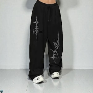 May include: Black sweatpants with a drawstring waist and white graphic designs. The pants have a loose, relaxed fit. The graphic designs are symmetrical and appear to be a stylized starburst or floral pattern.