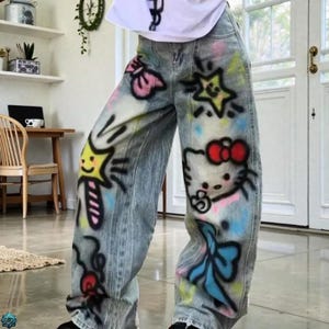 Pode incluir: Jeans de ganga de lavagem clara com desenhos coloridos em estilo grafite. Os jeans apresentam estrelas de desenhos animados, laços e um gráfico Hello Kitty. A obra de arte é em tons de rosa, azul, amarelo e preto, criando um visual lúdico e único.