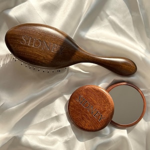 以下が含まれることがあります： 木製のヘアブラシと丸いコンパクトミラー。どちらも濃い色の木材で作られており、「SIDNEY」という名前が刻まれています。 ミラーは開いており、反射面が見えます。