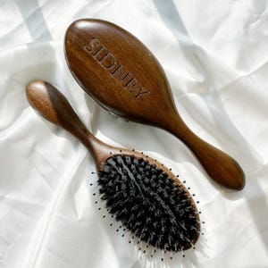 以下が含まれることがあります： 暗い毛と湾曲したハンドルが付いた2つの木製ヘアブラシ。大きなブラシの背面には「SIDNEY」という名前が刻まれています。ブラシは濃い茶色で、白くテクスチャ加工された表面に置かれています。