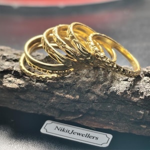Op de afbeelding: Een verzameling goudkleurige ringen met verschillende gegraveerde patronen, waaronder bloemen- en textuurontwerpen. De ringen worden gepresenteerd op een stuk donker hout, met een label met de tekst "NikitJewellers".
