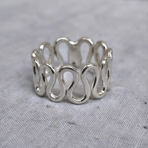 May include: A silver ring featuring a continuous, repeating wave-like pattern that forms an open, interconnected structure. The metal has a polished, reflective finish, creating a series of rounded loops that wrap around the finger.
