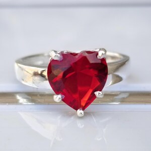 May include: A silver ring featuring a large, heart-shaped, faceted, deep red gemstone. The gemstone is securely held in place by four silver prongs. The band of the ring is a simple, polished silver.