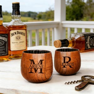 May include: Two stemless wooden whiskey glasses with personalized initials and names, alongside bottles of whiskey. The glasses are brown with black interiors. A corkscrew is visible.