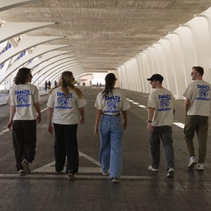 May include: Five people walking away from the camera, wearing cream-colored t-shirts with a blue graphic and the words "SMASH PATRIARCHY". They are walking down a wide, covered walkway with a modern architectural design.