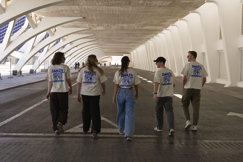 May include: Five people walking away from the camera, wearing cream-colored t-shirts with a blue graphic and the words "SMASH PATRIARCHY". They are walking down a wide, covered walkway with a modern architectural design.