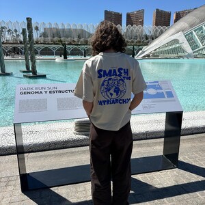 May include: A person wearing a cream-colored t-shirt with the text "SMASH the PATRIARCHY" in blue. The person is standing in front of a large body of water with modern architecture in the background. Brown pants complete the outfit.
