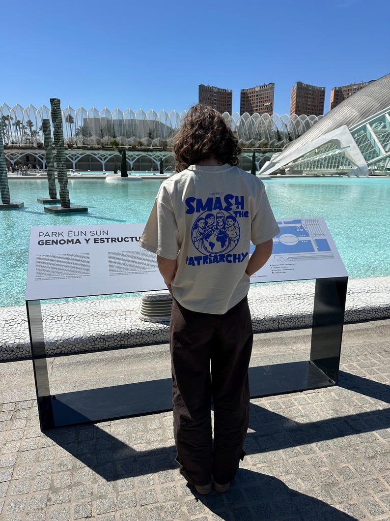May include: A person wearing a cream-colored t-shirt with the text "SMASH the PATRIARCHY" in blue. The person is standing in front of a large body of water with modern architecture in the background. Brown pants complete the outfit.