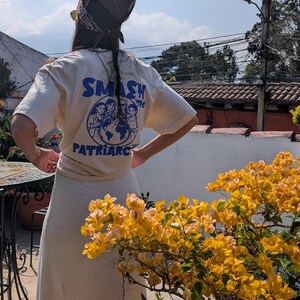 May include: A person wearing a cream-colored t-shirt with the words "SMASH THE PATRIARCHY" in blue, along with a graphic of women around the globe. The person is also wearing a brown bandana and a cream-colored skirt.