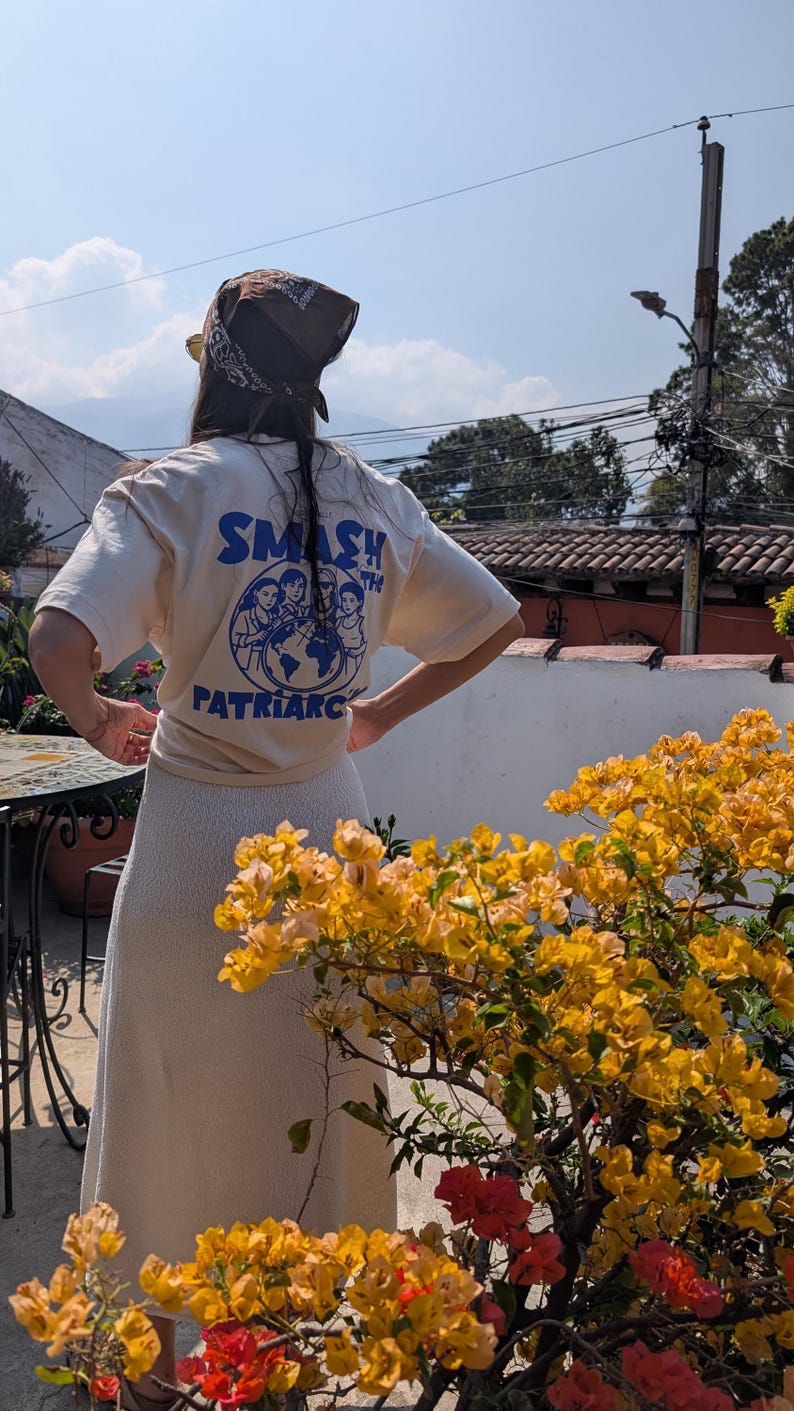 May include: A person wearing a cream-colored t-shirt with the words "SMASH THE PATRIARCHY" in blue, along with a graphic of women around the globe. The person is also wearing a brown bandana and a cream-colored skirt.
