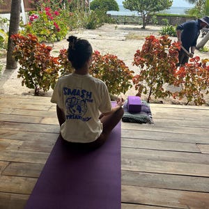 May include: A person is seated on a purple yoga mat, practicing yoga outdoors. The person is wearing a cream-colored t-shirt with the text "SMASH the TRIARCHY" printed on the back. A yoga block and a rolled-up mat are nearby.