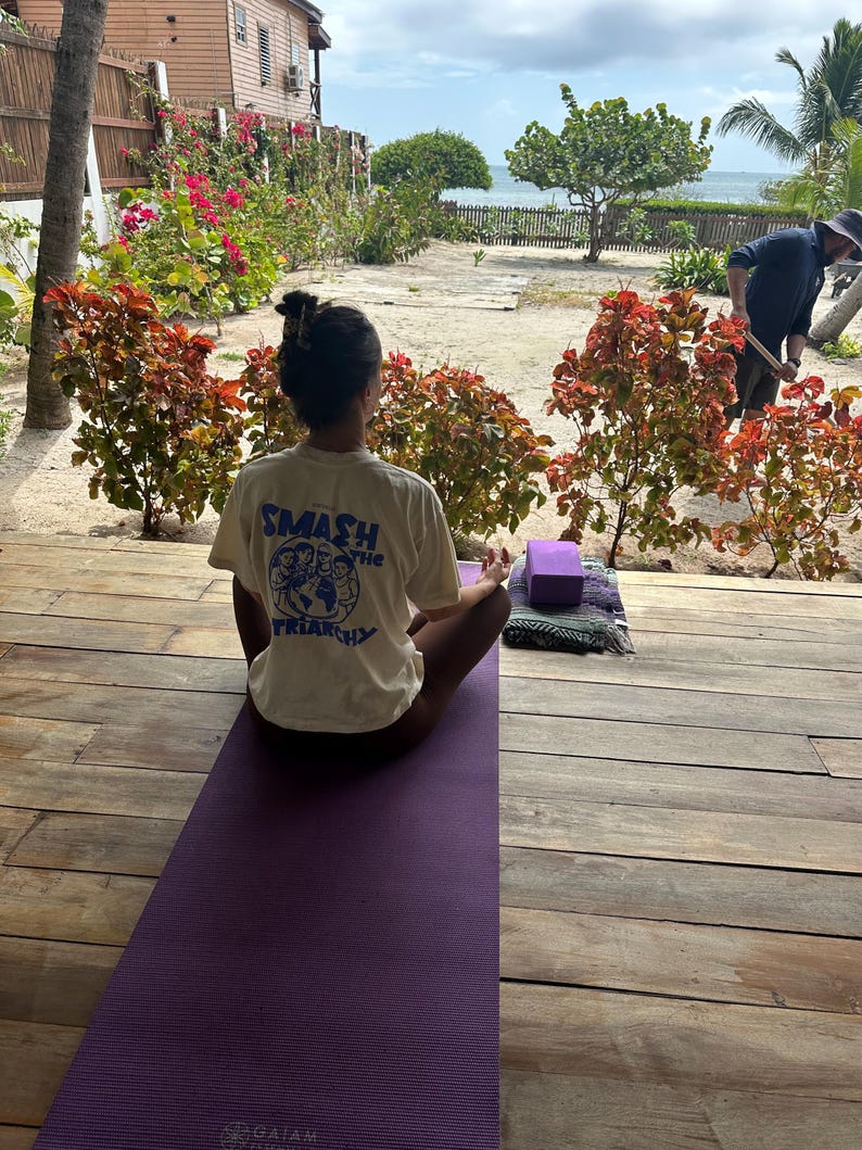 May include: A person is seated on a purple yoga mat, practicing yoga outdoors. The person is wearing a cream-colored t-shirt with the text "SMASH the TRIARCHY" printed on the back. A yoga block and a rolled-up mat are nearby.