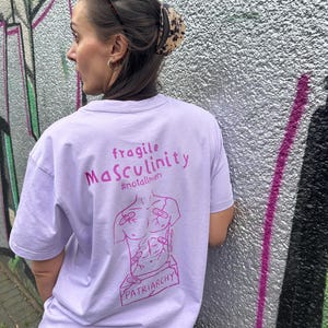 Könnte beinhalten: Ein hellviolettes T-Shirt mit dunkelrosa Text, der fragile MaSculinity #notallmen lautet. Unter dem Text befindet sich eine Strichzeichnung eines Torsos mit Pflastern und dem Wort PATRIARCHY, das auf einer Basis am unteren Rand gedruckt ist.