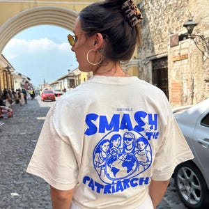 Może przedstawiać: Kremowa koszulka z niebieskim napisem "SMASH the PATRIARCHY" oraz grafiką kobiet otaczających globus. Na koszulce znajduje się również napis "SOSTENELLE".