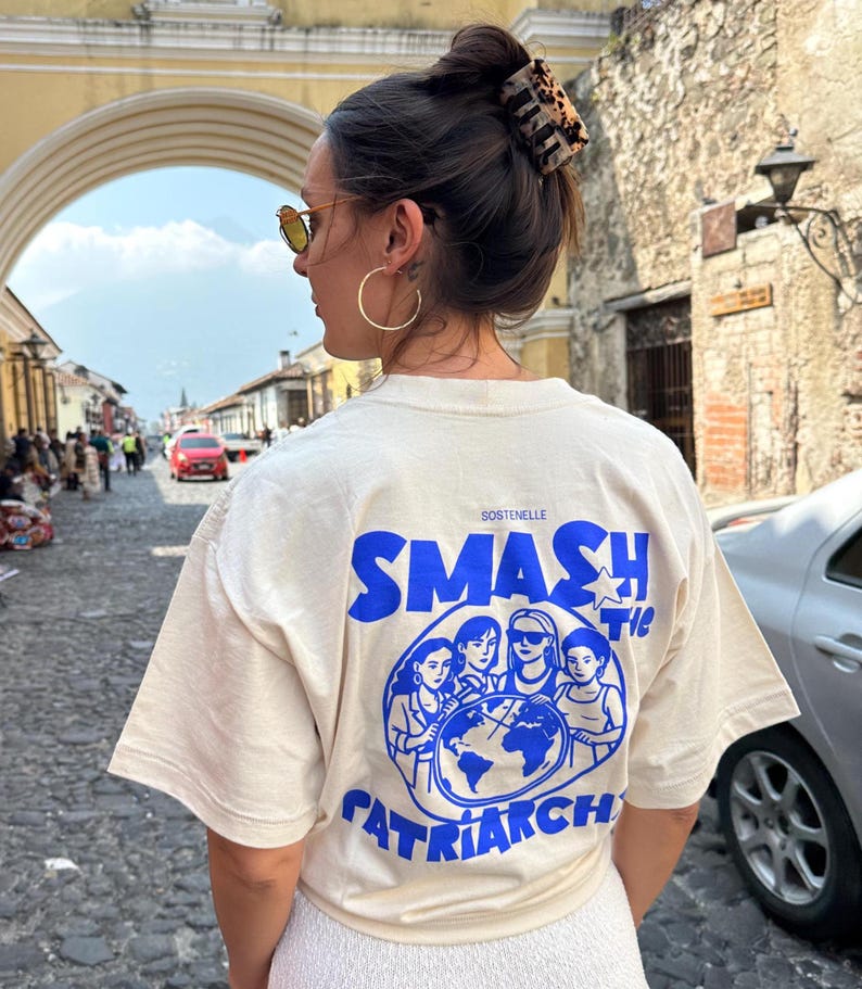 May include: Cream-colored t-shirt with the text "SMASH the PATRIARCHY" in blue, along with a graphic of women surrounding a globe. The shirt also has the word "SOSTENELLE" printed on it.