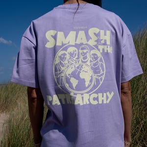 Könnte beinhalten: Ein hellviolettes Kurzarm-T-Shirt mit einer blassgelben Grafik auf der Rückseite. Das Design zeigt vier Figuren, die einen Globus umgeben, mit dem Text SMASH THE PATRIARCHY und SOSTENELLE in einer fetten, stilisierten Schriftart darüber und darunter.
