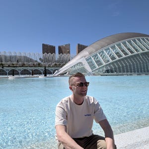 May include: A person wearing sunglasses and a cream-colored t-shirt with the text "InTheThrd" in blue, sitting near a pool of water. The background features modern architecture and a clear blue sky.