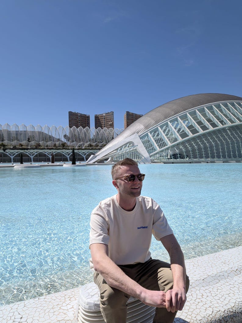 May include: A person wearing sunglasses and a cream-colored t-shirt with the text "InTheThrd" in blue, sitting near a pool of water. The background features modern architecture and a clear blue sky.