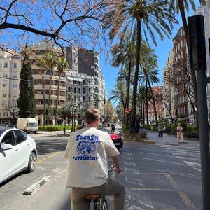 May include: A person riding a bicycle down a city street, wearing a cream-colored t-shirt with the text "SMASH THE PATRIARCHY" in blue. The image is taken from behind, showing the person's back. The background includes palm trees, buildings, and a clear blue sky.
