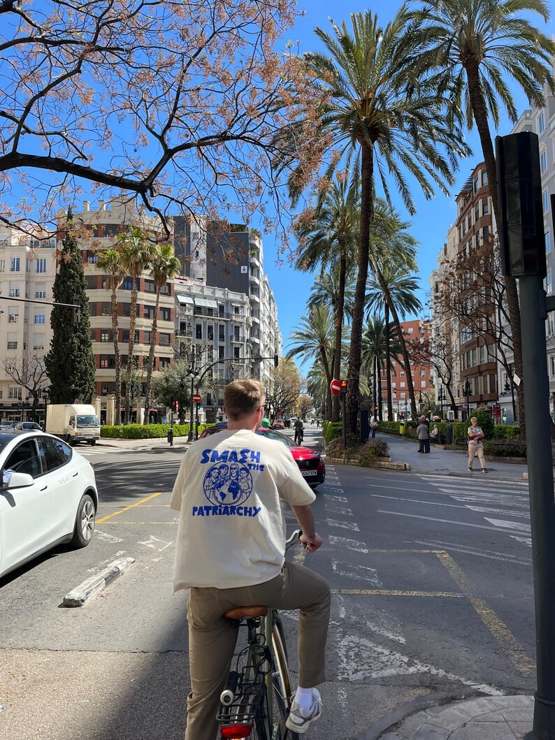 May include: A person riding a bicycle down a city street, wearing a cream-colored t-shirt with the text "SMASH THE PATRIARCHY" in blue. The image is taken from behind, showing the person's back. The background includes palm trees, buildings, and a clear blue sky.