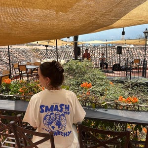 May include: A person wearing a cream-colored t-shirt with the words "SMASH THE ARCHY" in blue, along with a graphic of people and a globe. The person is sitting at a table on a rooftop patio with orange flowers and a band playing in the background.
