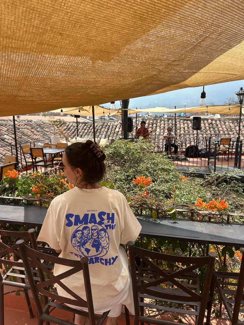 May include: A person wearing a cream-colored t-shirt with the words "SMASH THE ARCHY" in blue, along with a graphic of people and a globe. The person is sitting at a table on a rooftop patio with orange flowers and a band playing in the background.
