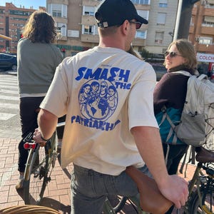 May include: A person wearing a cream-colored t-shirt with the text "SMASH THE PATRIARCHY" in blue. The shirt features a graphic of several women and a globe. The person is riding a bicycle.
