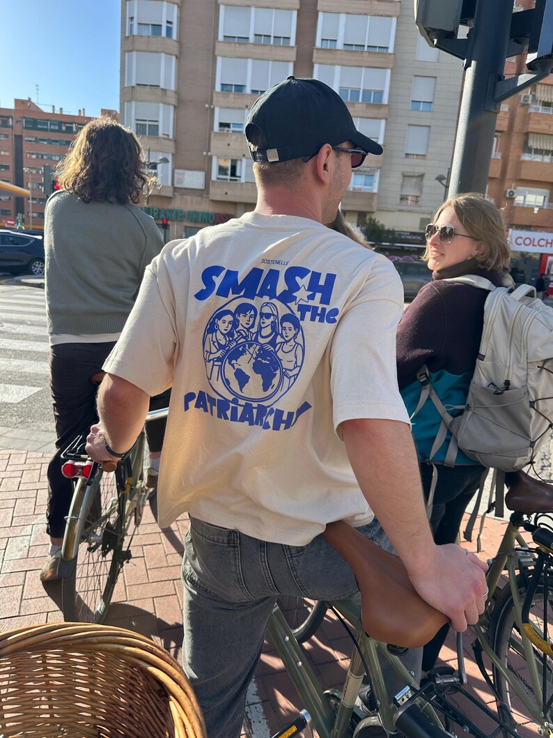 May include: A person wearing a cream-colored t-shirt with the text "SMASH THE PATRIARCHY" in blue. The shirt features a graphic of several women and a globe. The person is riding a bicycle.