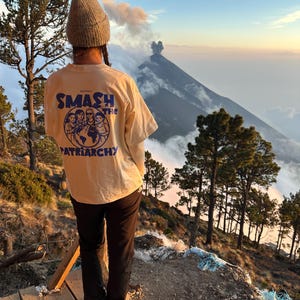 May include: A person wearing a light yellow t-shirt with the words "SMASH THE PATRIARCHY" in blue and white. The person is standing on a wooden platform, with a mountain and trees in the background. The sky is a mix of blue and orange.