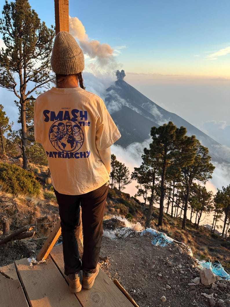 May include: A person wearing a light yellow t-shirt with the words "SMASH THE PATRIARCHY" in blue and white. The person is standing on a wooden platform, with a mountain and trees in the background. The sky is a mix of blue and orange.