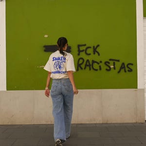 May include: A person wearing a cream-colored t-shirt with a blue graphic and the words "SMASH PATRIARCHY" on the back, paired with light blue jeans. The background features green wall with graffiti that reads "FCK RACISTAS".