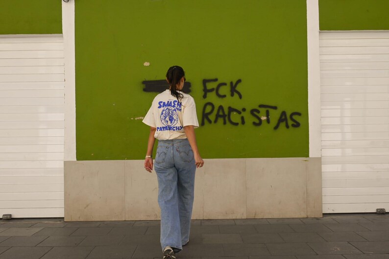 May include: A person wearing a cream-colored t-shirt with a blue graphic and the words "SMASH PATRIARCHY" on the back, paired with light blue jeans. The background features green wall with graffiti that reads "FCK RACISTAS".