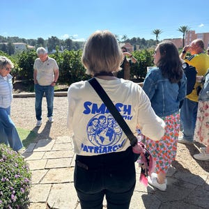 May include: A cream-colored t-shirt with the words "SMASH THE PATRIARCHY" in blue, featuring a graphic of people around a globe. The shirt is worn by a person standing outdoors with other people in the background.