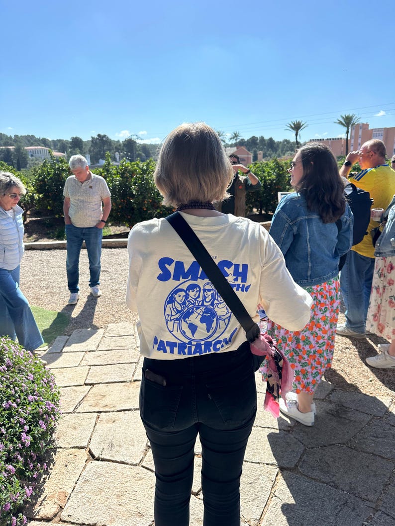 May include: A cream-colored t-shirt with the words "SMASH THE PATRIARCHY" in blue, featuring a graphic of people around a globe. The shirt is worn by a person standing outdoors with other people in the background.