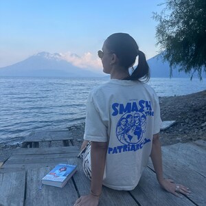 May include: A cream-colored t-shirt with the text "SMASH THE PATRIARCHY" in blue, featuring a graphic of people around a globe. A book with a blue cover is visible. The scene is set outdoors with a mountain and water in the background.