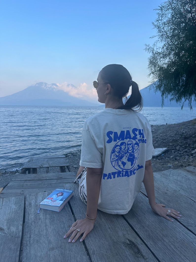 May include: A cream-colored t-shirt with the text "SMASH THE PATRIARCHY" in blue, featuring a graphic of people around a globe. A book with a blue cover is visible. The scene is set outdoors with a mountain and water in the background.