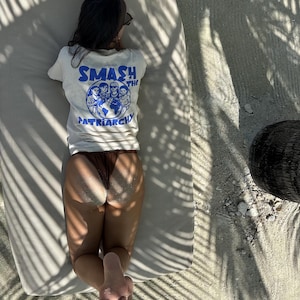 May include: A person wearing a white t-shirt with the text "SMASH THE PATRIARCHY" in blue, lying on a beach chair. The shirt features a graphic of people around a globe. Brown bottoms and sand are also visible.