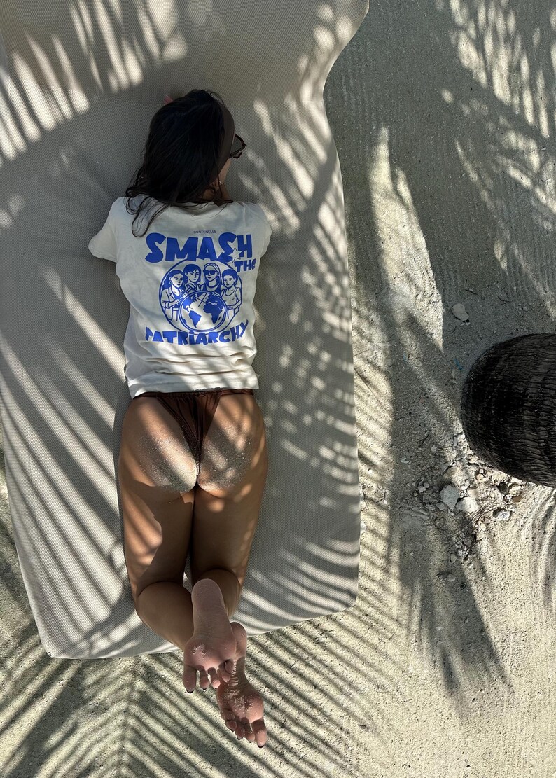 May include: A person wearing a white t-shirt with the text "SMASH THE PATRIARCHY" in blue, lying on a beach chair. The shirt features a graphic of people around a globe. Brown bottoms and sand are also visible.