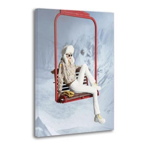 May include: A canvas print depicts a person seated on a red ski lift chair against a snowy mountain backdrop. The person wears a zebra-print jacket, white pants, a fur hat, sunglasses, and white sneakers. Yellow gloves rest on the seat.