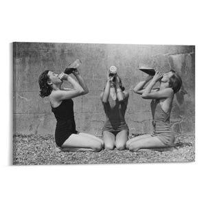 May include: Black and white photograph of three people kneeling and drinking from bottles. They are wearing vintage swimsuits and are positioned in front of a textured wall. The image has a retro, nostalgic feel.