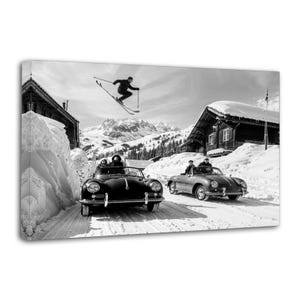 May include: Black and white canvas print of a vintage ski scene. A skier is mid-air above two classic convertible cars parked on a snow-covered road. Wooden chalets and snow-capped mountains are in the background.